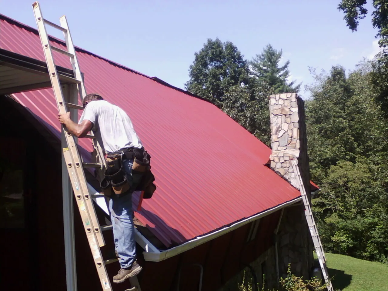 Roofer on a ladder beside a red metal roof and stone chimney — Soffit & Fascia Repair by Roofing Brothers in Hankamer